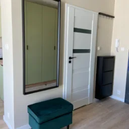 Nowoczesny przedpokój z lustrem w czarnej ramie, zielonym pufem i białą, panelową ścianą z drzwiami, uzupełniony czarną szafką na buty i wieszakiem ściennym.