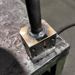 Wojciech Czerenda - Metalowy sześcian z przyspawaną rurą, widoczne ślady spawania i otwory montażowe, całość umieszczona na metalowym stole warsztatowym.