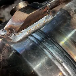 Spoina na metalowej rurze, wykonana metodą punktową, widoczne ślady obróbki i niewielkie krople na powierzchni.