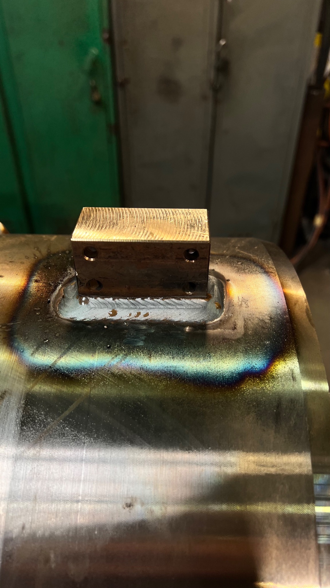 Metalowy blok z otworami przymocowany do obrobionego elementu z widocznymi śladami spawania i opalizującymi przebarwieniami.