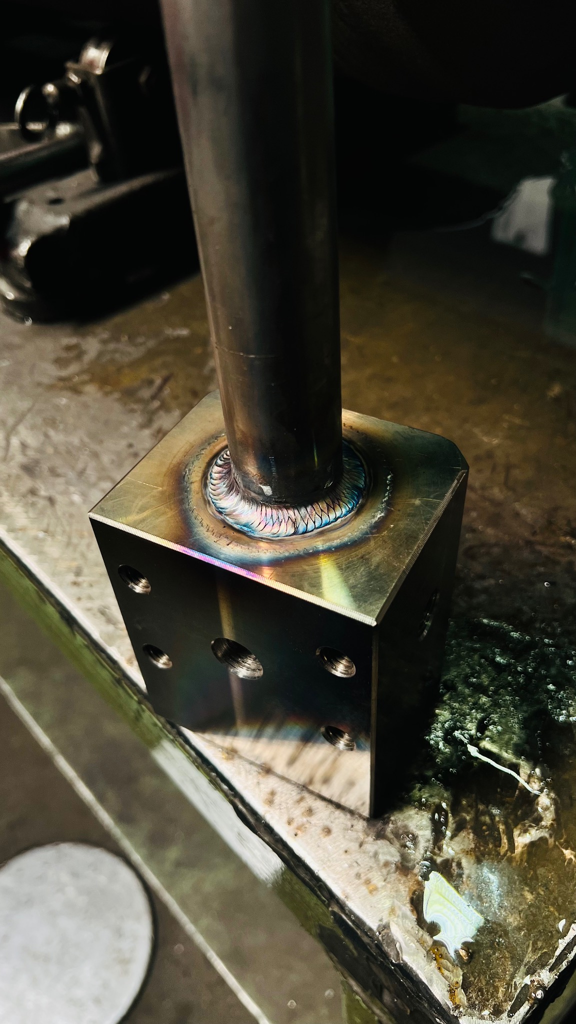 Metalowy sześcian z przyspawaną rurą, widoczne ślady spawania TIG z charakterystycznymi kolorami termicznymi na spoinie, otoczenie warsztatowe.