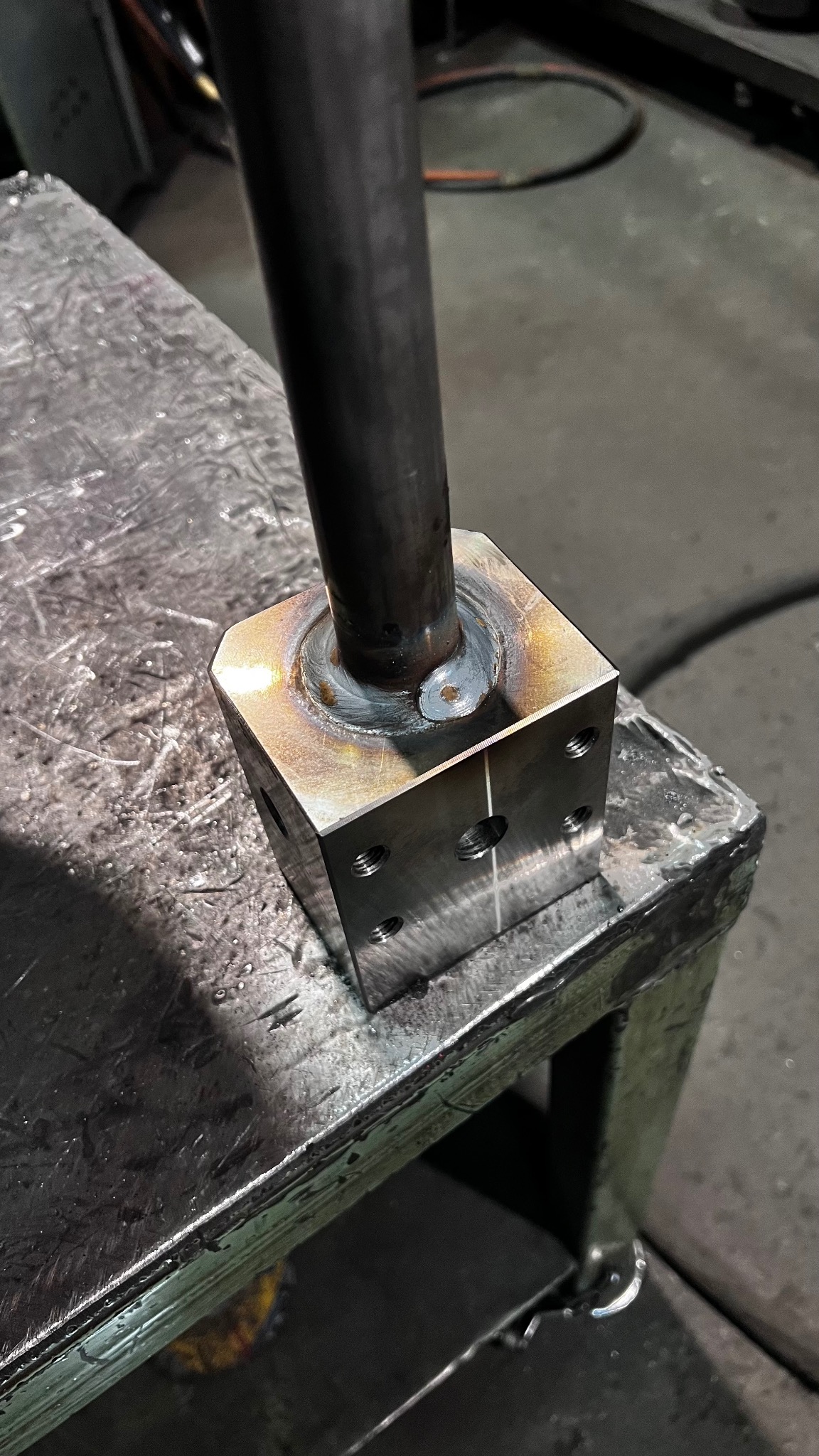 Metalowy sześcian z przyspawaną rurą, widoczne ślady spawania i otwory montażowe, całość umieszczona na metalowym stole warsztatowym.