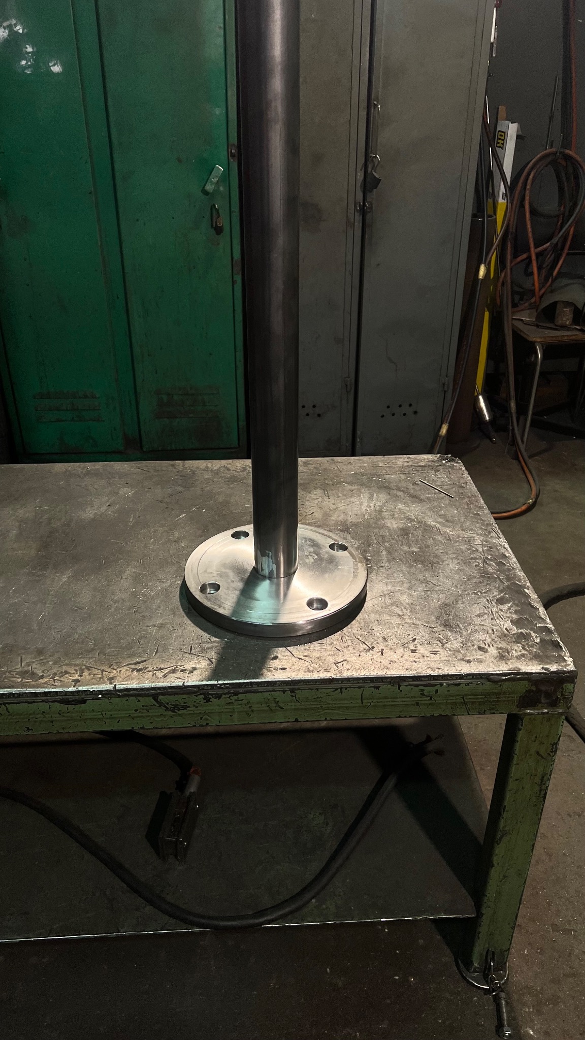 Metalowy słup przymocowany do okrągłej podstawy z otworami na śruby, ustawiony na roboczym stole w warsztacie, z zielonymi szafkami w tle.