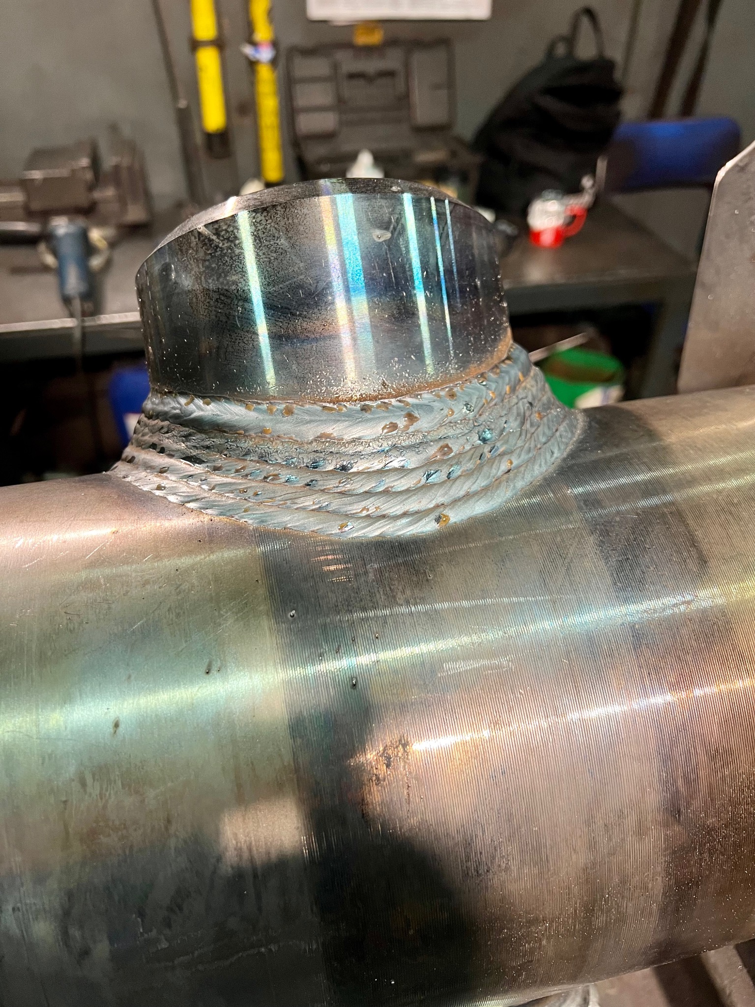 Spoina pierścieniowa na metalowej rurze, widoczne ślady po procesie obróbki, z efektem tęczy na powierzchni. W tle warsztat.