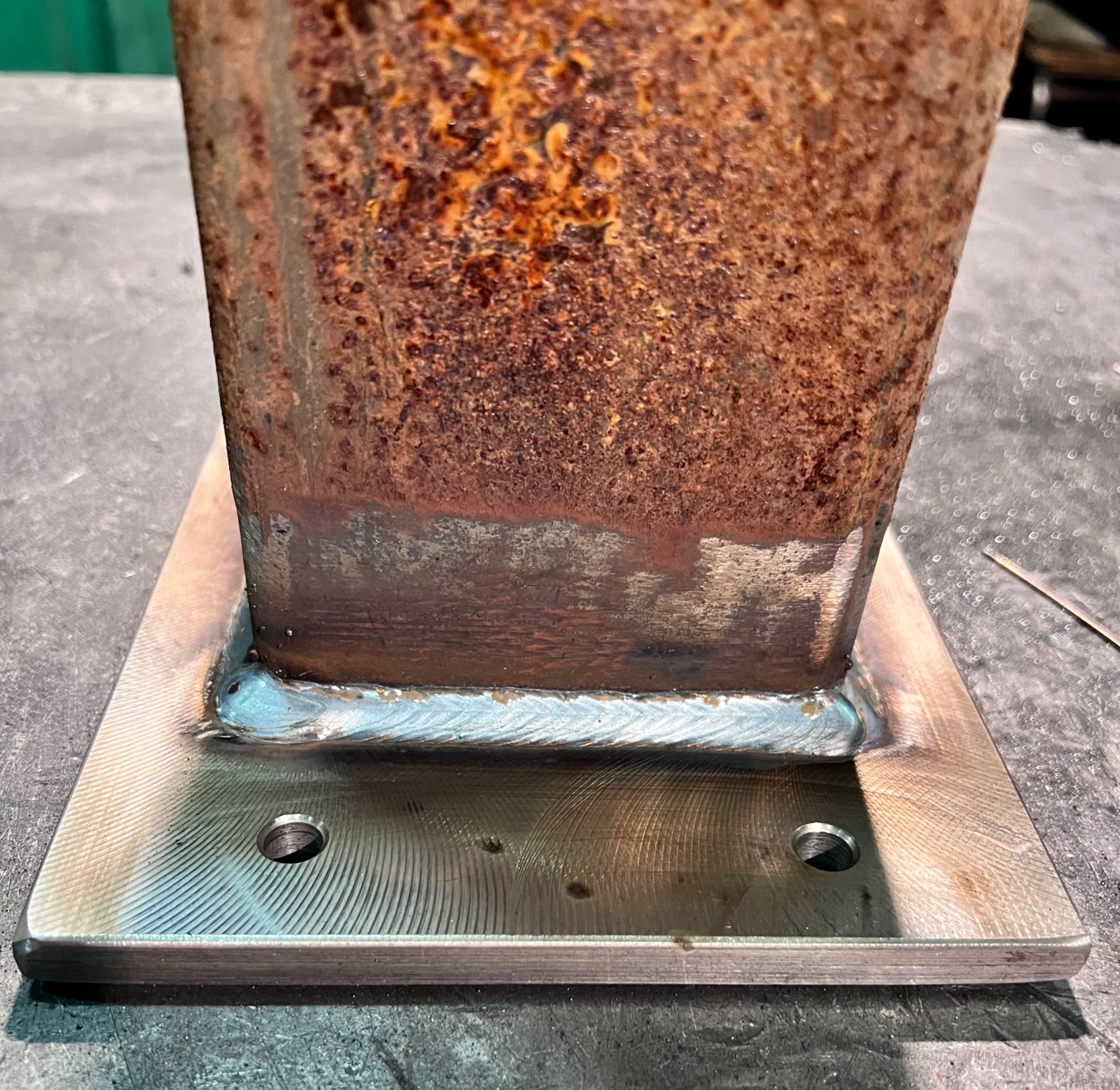 Zespawany, rdzawy profil kwadratowy przymocowany do metalowej płyty z widocznymi otworami na śruby.