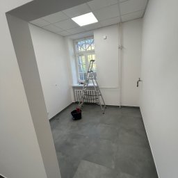 Przedsiębiorstwo Budowlane Solid-Max - Wnętrze pokoju po malowaniu ścian na biało, widoczna drabina oparta o okno, wiadro z farbą i szary gres na podłodze.