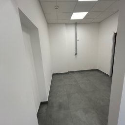 Przedsiębiorstwo Budowlane Solid-Max - Puste pomieszczenie po remoncie z szarą podłogą z płytek, białymi ścianami i sufitem kasetonowym z wbudowanymi lampami, widoczny fragment instalacji na ścianie i ościeżnica drzwiowa.