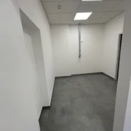 Puste pomieszczenie po remoncie z szarą podłogą z płytek, białymi ścianami i sufitem kasetonowym z wbudowanymi lampami, widoczny fragment instalacji na ścianie i ościeżnica drzwiowa.