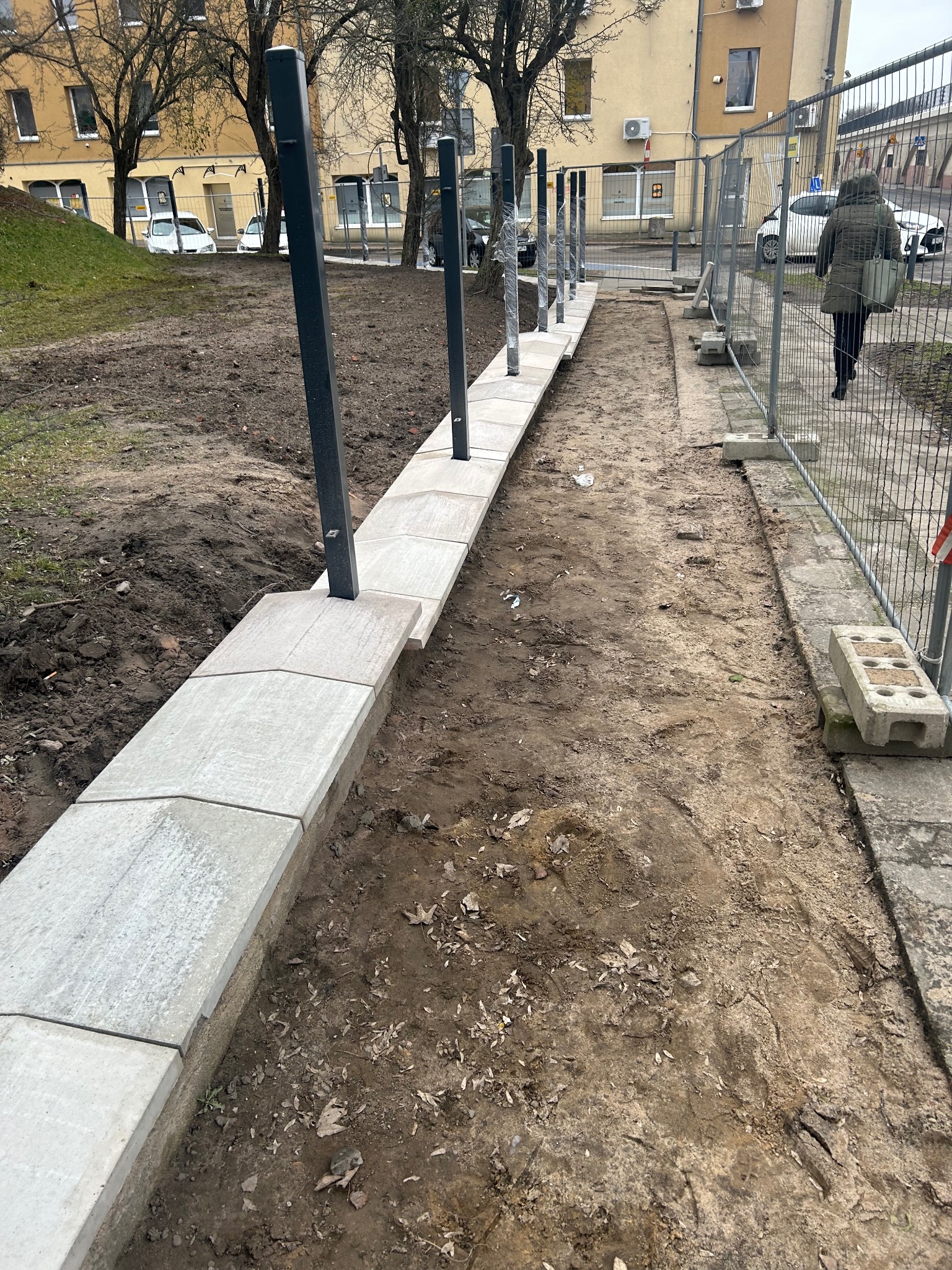 Świeżo ułożony, niski murek z szarych, betonowych płyt z zamontowanymi, czarnymi słupkami oświetleniowymi, teren wokół w stanie surowym, widoczne ślady prac ziemnych, w tle budynek mieszkalny...