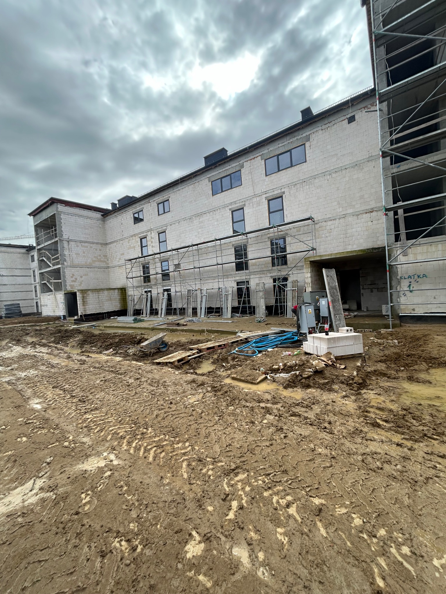 Budynek w trakcie budowy z białych bloków, otoczony rusztowaniami i błotnistym terenem w pochmurny dzień.
