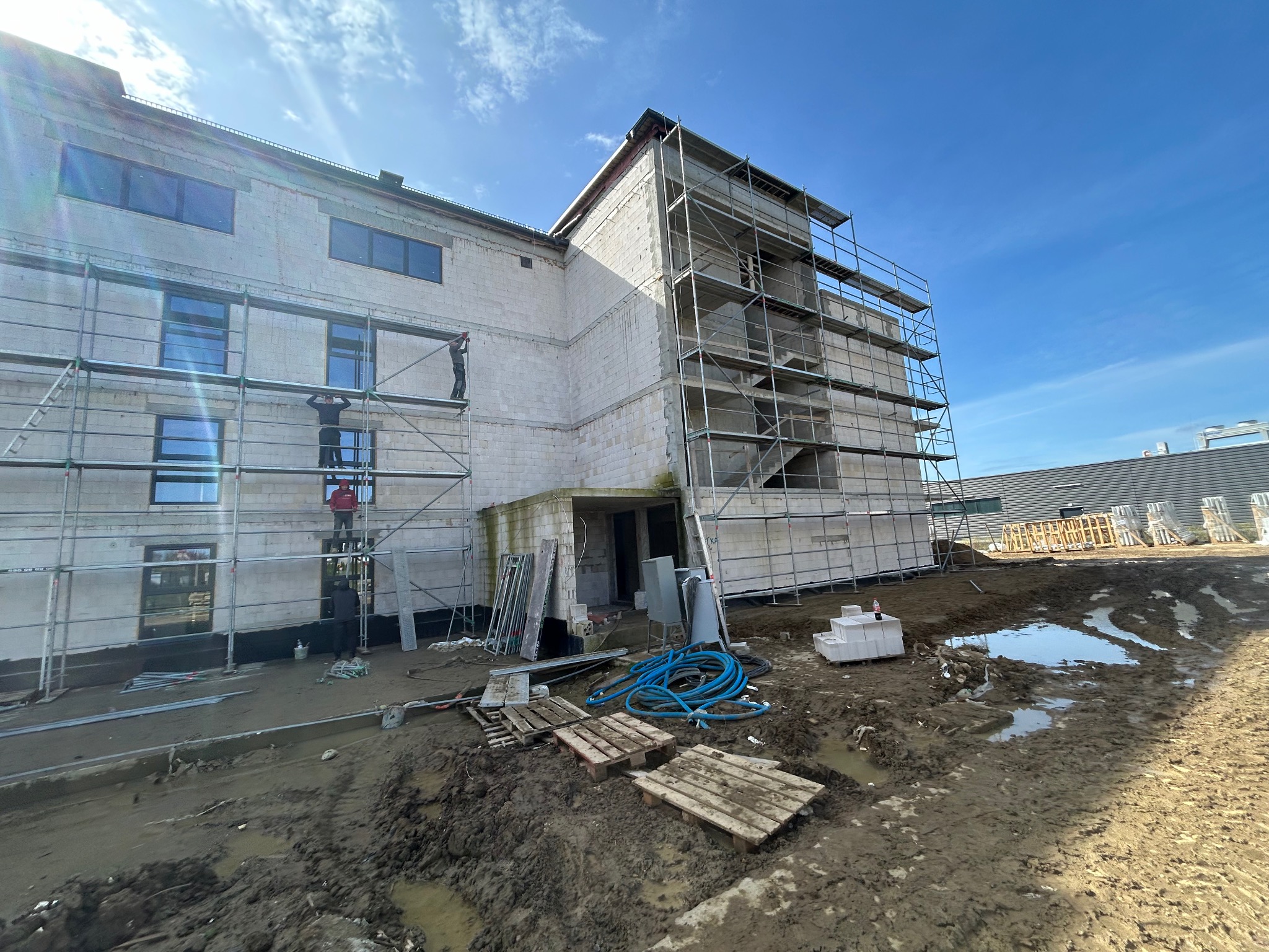 Budynek w trakcie ocieplania, z widocznymi rusztowaniami, materiałami budowlanymi i pracownikami na elewacji, błotniste podłoże.