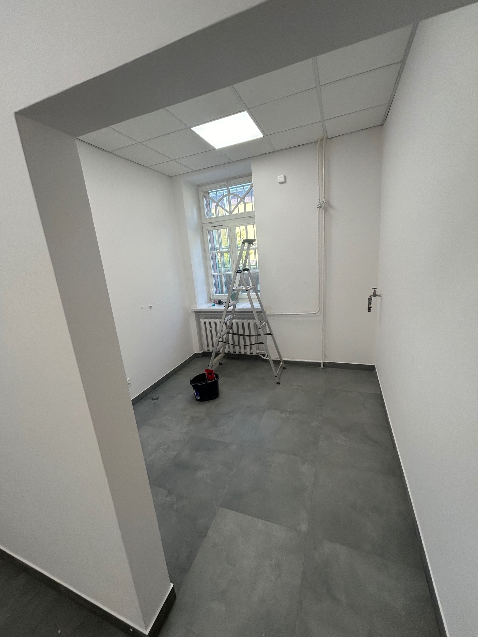 Wnętrze pokoju po malowaniu ścian na biało, widoczna drabina oparta o okno, wiadro z farbą i szary gres na podłodze.