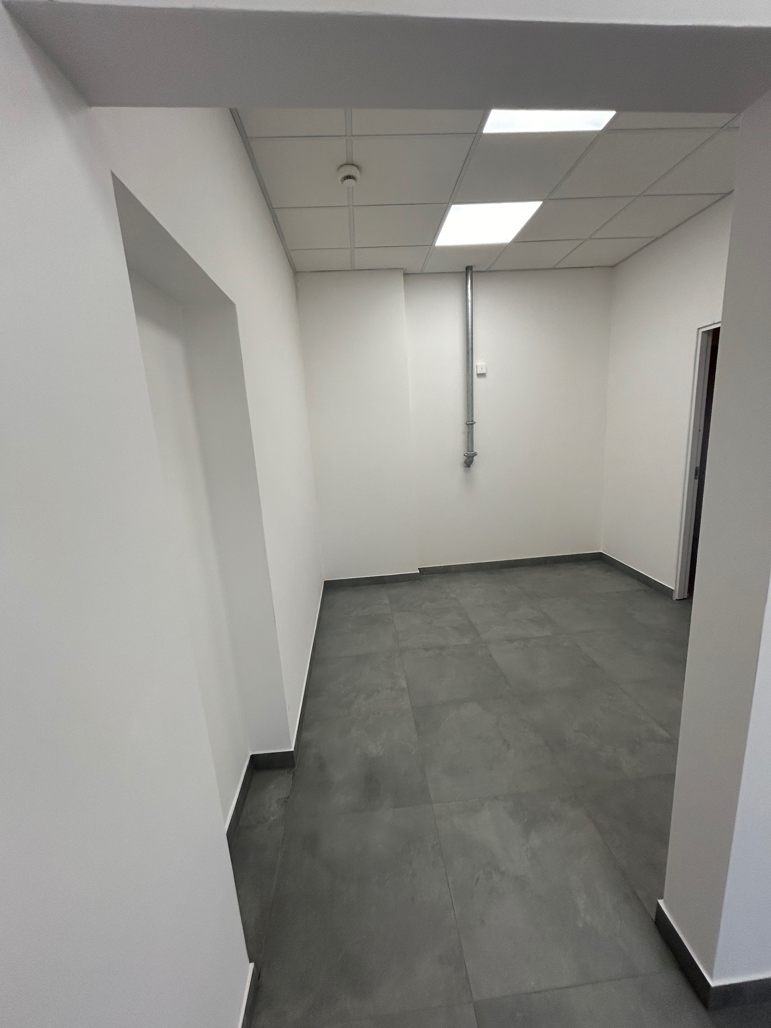 Puste pomieszczenie po remoncie z szarą podłogą z płytek, białymi ścianami i sufitem kasetonowym z wbudowanymi lampami, widoczny fragment instalacji na ścianie i ościeżnica drzwiowa.