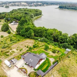 Panoramiczny widok z drona na dom z ciemną dachówką, panele solarne na dachu, dwa samochody i busa zaparkowane na podjeździe, otoczony zielenią i jeziorem w tle.