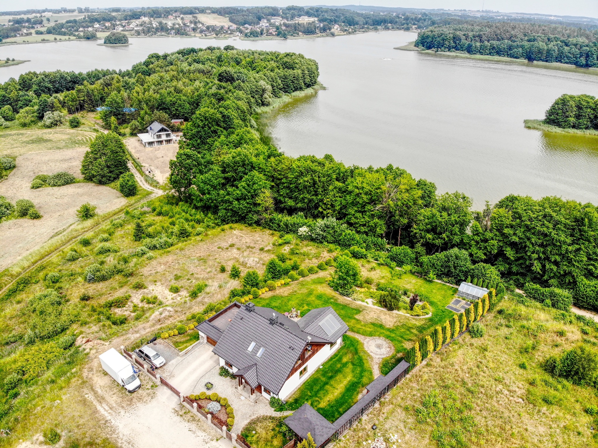 Panoramiczny widok z drona na dom z ciemną dachówką, panele solarne na dachu, dwa samochody i busa zaparkowane na podjeździe, otoczony zielenią i jeziorem w tle.