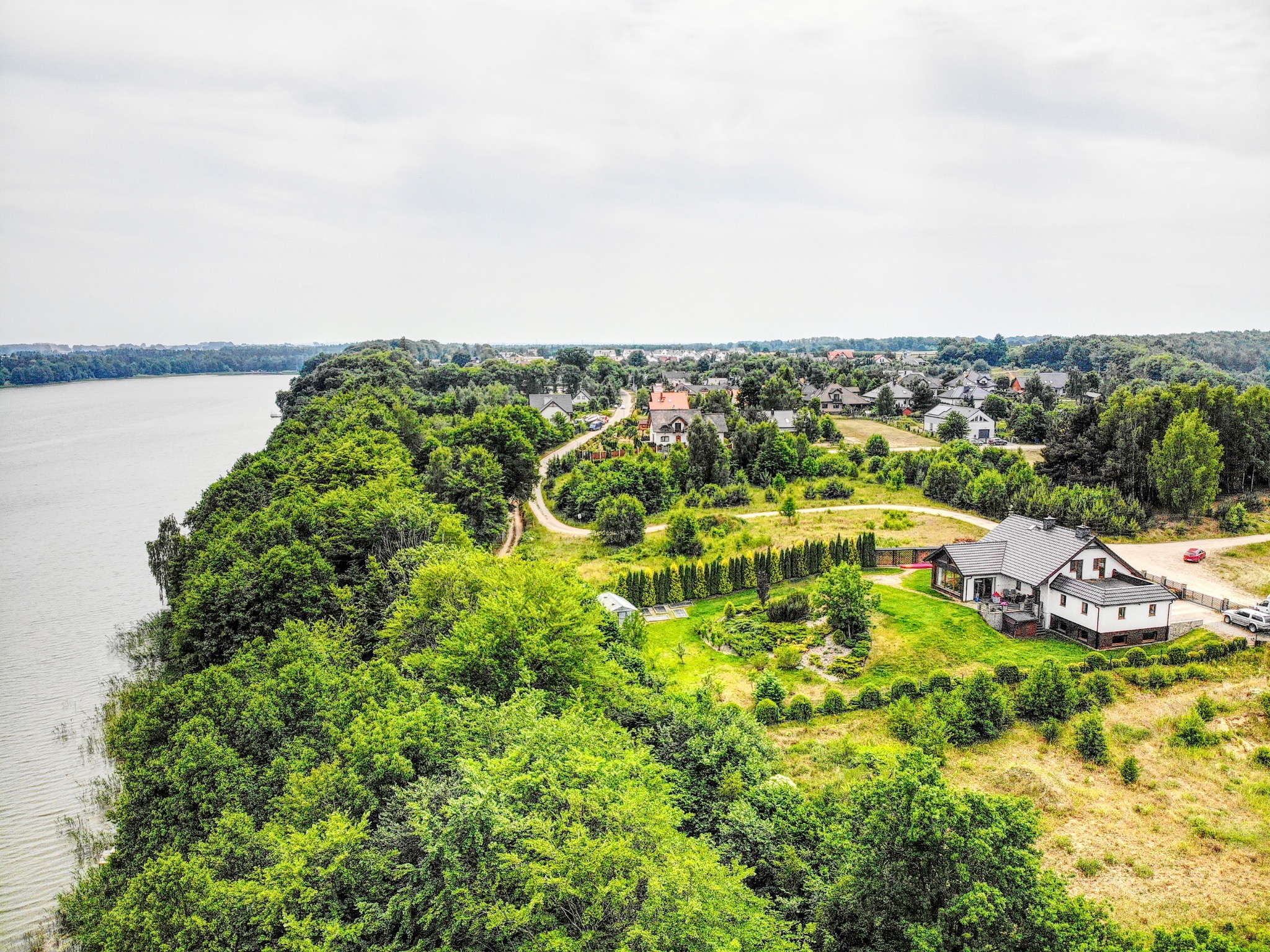 Panoramiczny widok z lotu ptaka na dom jednorodzinny z ogrodem, położony nad brzegiem jeziora, otoczony gęstą zielenią drzew i zabudową wiejską w oddali.