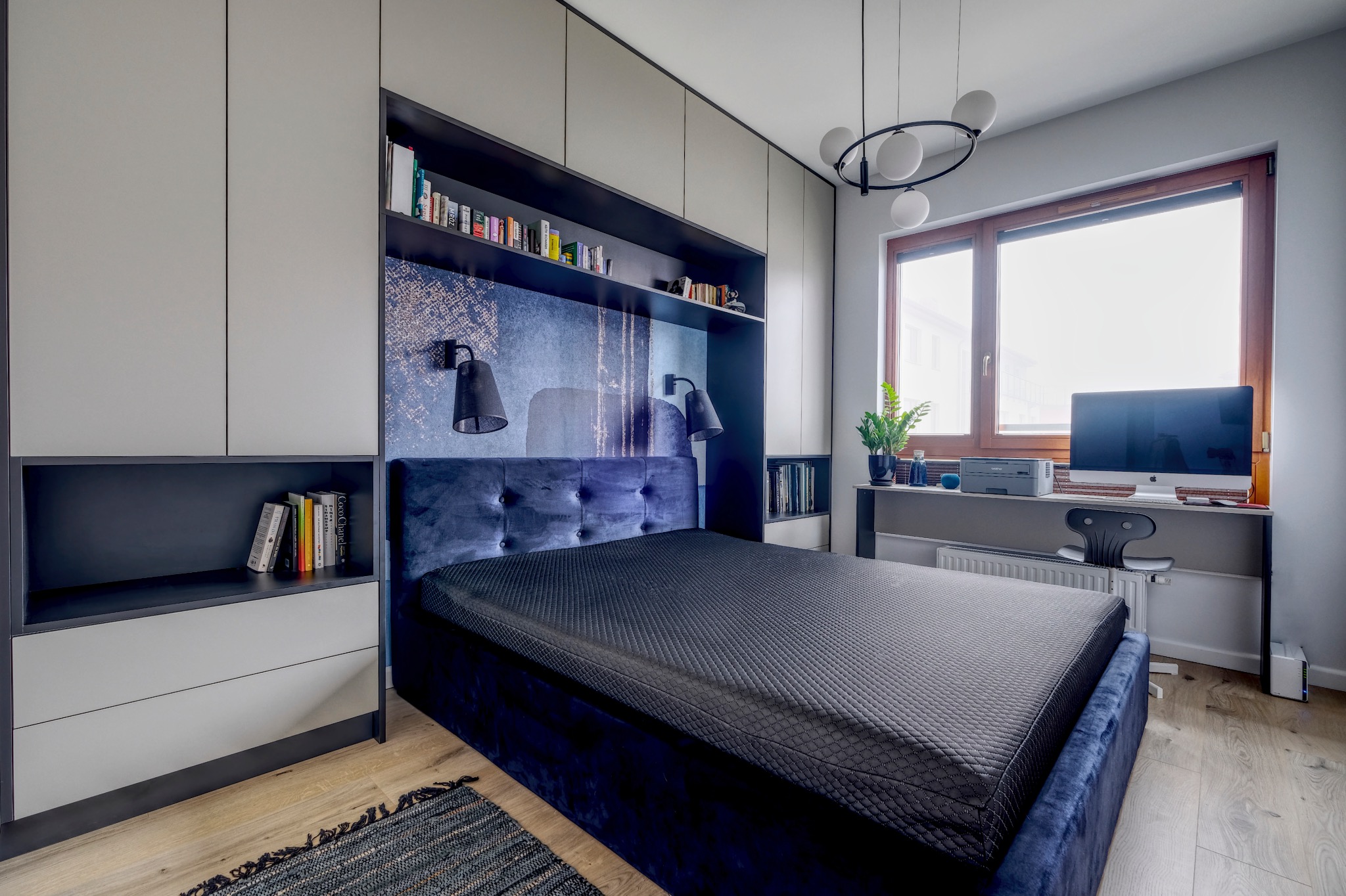 Nowoczesna sypialnia z łóżkiem z ciemnoniebieskim wezgłowiem i czarną narzutą, wbudowanymi szafami z półkami na książki i biurkiem z komputerem przy oknie.