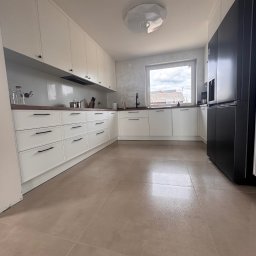 B&B Company - Jasna kuchnia z białymi szafkami, brązowym blatem i dużymi, beżowymi płytkami podłogowymi. Widoczny czarny, nowoczesny, dwudrzwiowy lodówka.