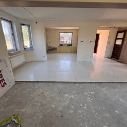 B&B Company - Wnętrze w trakcie układania białych płytek gresowych na podłodze. Widoczne okna, grzejnik, karton z napisem 'GRES' i siedząca osoba w oddali.