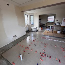 B&B Company - Układanie płytek podłogowych z użyciem klipsów poziomujących w przestronnym wnętrzu z oknami i przygotowaną ścianą. Widoczny sprzęt glazurniczy.