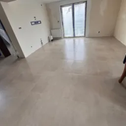 Jasne, matowe płytki podłogowe w przestronnym pokoju z oknem balkonowym i przygotowanymi instalacjami na ścianach. Widoczny fragment krzesła i zabezpieczone folią drzwi.