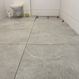 B&B Company - Szare płytki podłogowe w łazience, ułożone z użyciem systemu poziomowania. Widoczne płytki na ścianie i podwieszana toaleta z zabezpieczeniem.