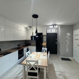 B&B Company - Nowoczesna kuchnia z białymi szafkami, drewnianym blatem i stołem jadalnym. Minimalistyczne oświetlenie i szara ściana z dekoracją kwiatową.