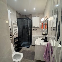 B&B Company - Nowoczesna łazienka w szarościach z kabiną prysznicową w czarnej ramie, białą armaturą i dekoracyjną ścianą z lustrem. Widok na całe wnętrze.