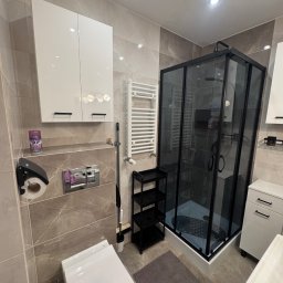 B&B Company - Wykończona łazienka w beżowych płytkach z kabiną prysznicową w czarnej ramie, toaletą i białymi szafkami. Grzejnik drabinkowy i czarne półki dodają funkcjonalności.