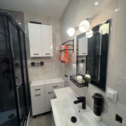 B&B Company - Nowoczesna łazienka po remoncie: białe szafki z czarnymi uchwytami, kabina prysznicowa z czarnymi ramami, szare płytki, czarna armatura i oświetlenie.