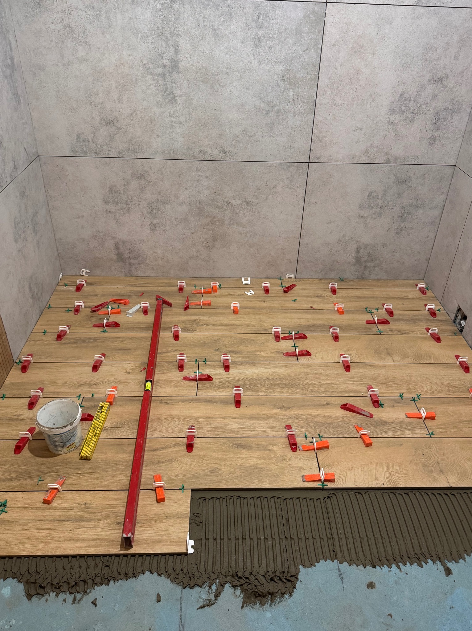 Układanie płytek imitujących drewno na podłodze w łazience. Widoczne narzędzia glazurnicze, poziomica i system poziomowania płytek. Ściany wyłożone płytkami w odcieniach szarości.