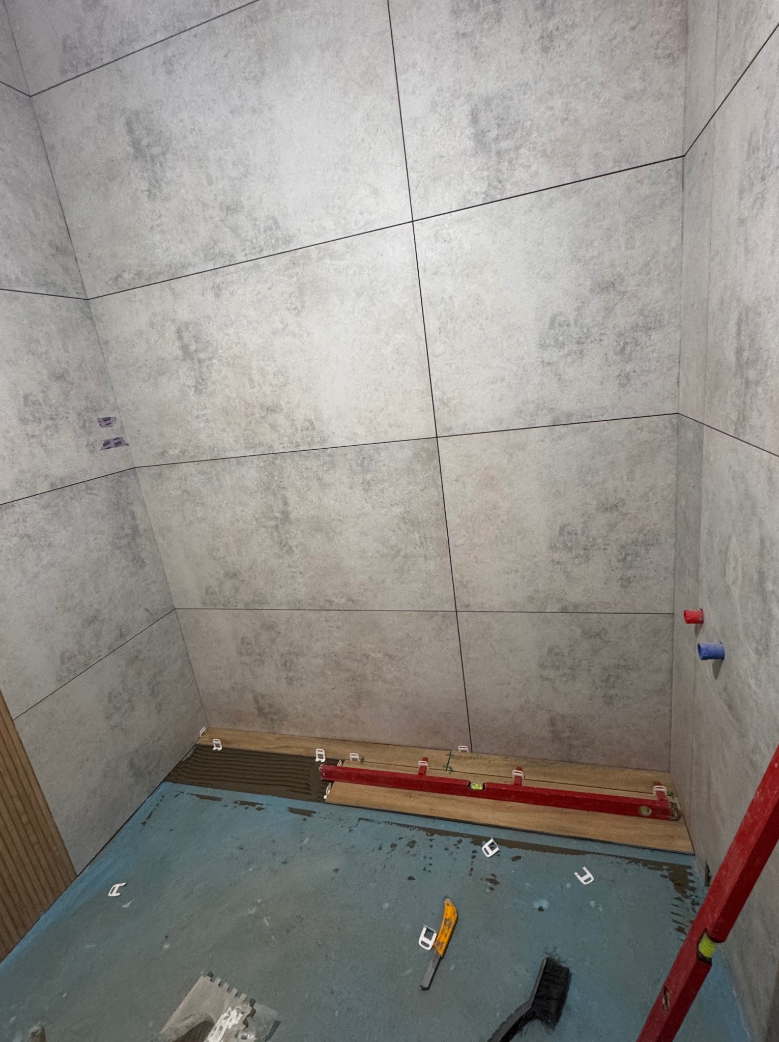 Łazienka w trakcie wykańczania: szare płytki na ścianach z fugą, poziomica i narzędzia glazurnicze na podłodze. Widoczne fragmenty instalacji wodnej.