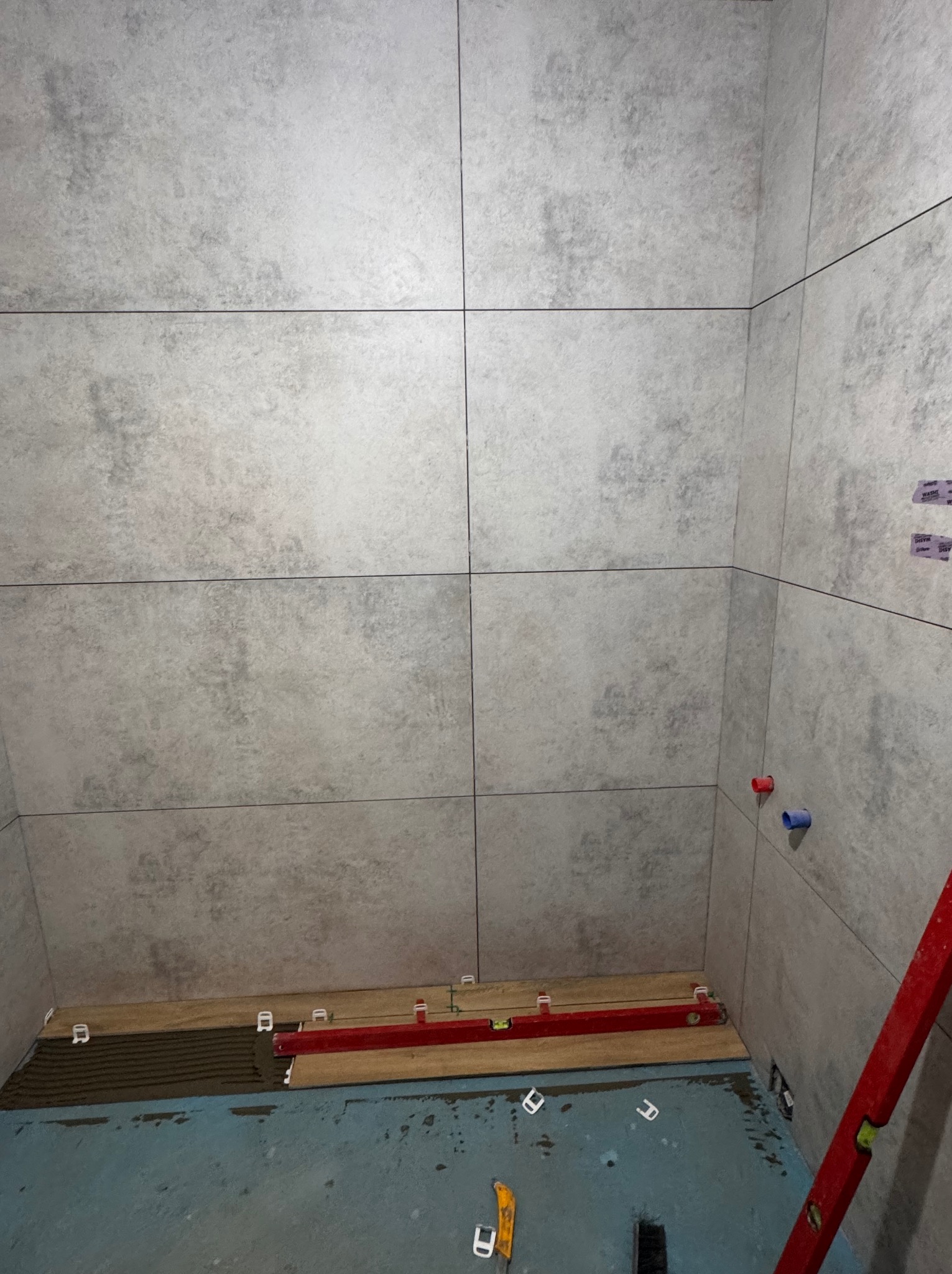 Wnętrze łazienki w trakcie prac glazurniczych. Na ścianach ułożone duże, szare płytki imitujące beton. Na podłodze poziomica i elementy do poziomowania płytek.
