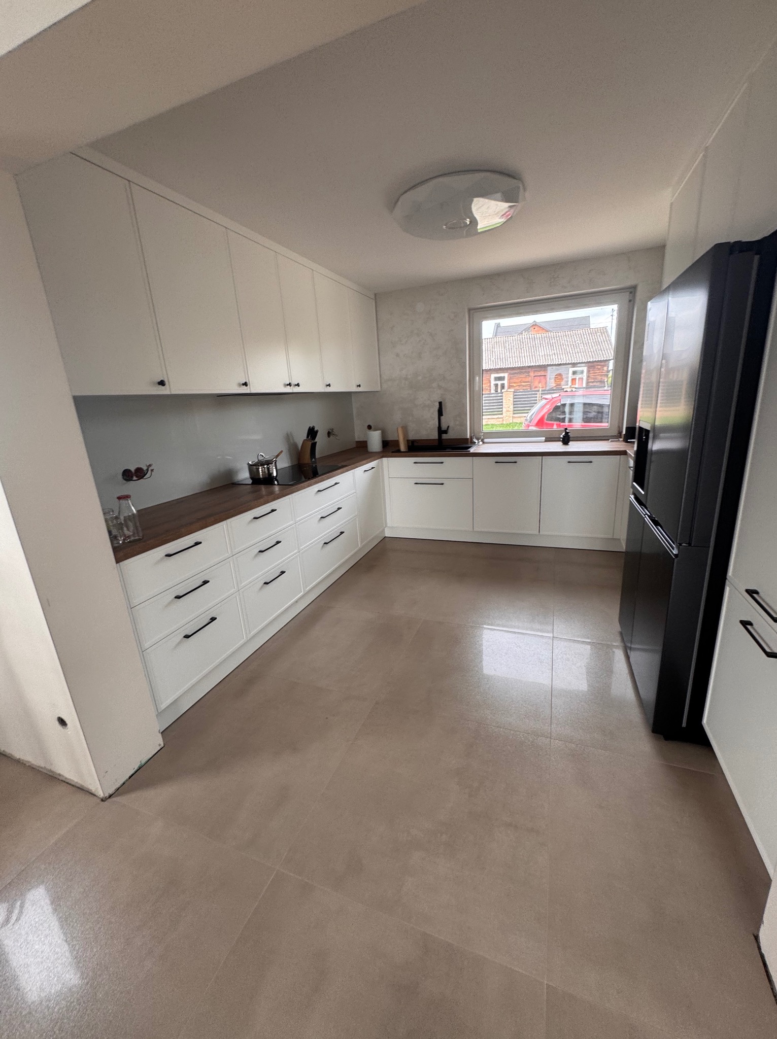 Nowoczesna, jasna kuchnia z białymi szafkami, drewnianym blatem i czarną lodówką. Widok na okno z zabudową. Minimalistyczny design i neutralna kolorystyka.