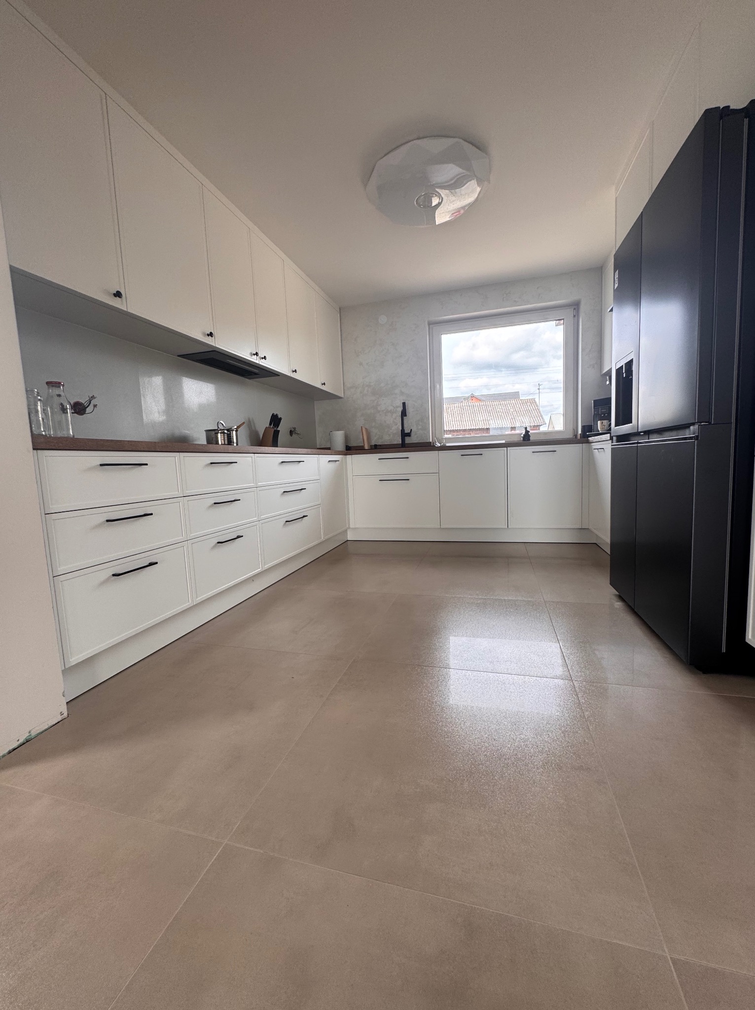 Jasna kuchnia z białymi szafkami, brązowym blatem i dużymi, beżowymi płytkami podłogowymi. Widoczny czarny, nowoczesny, dwudrzwiowy lodówka.