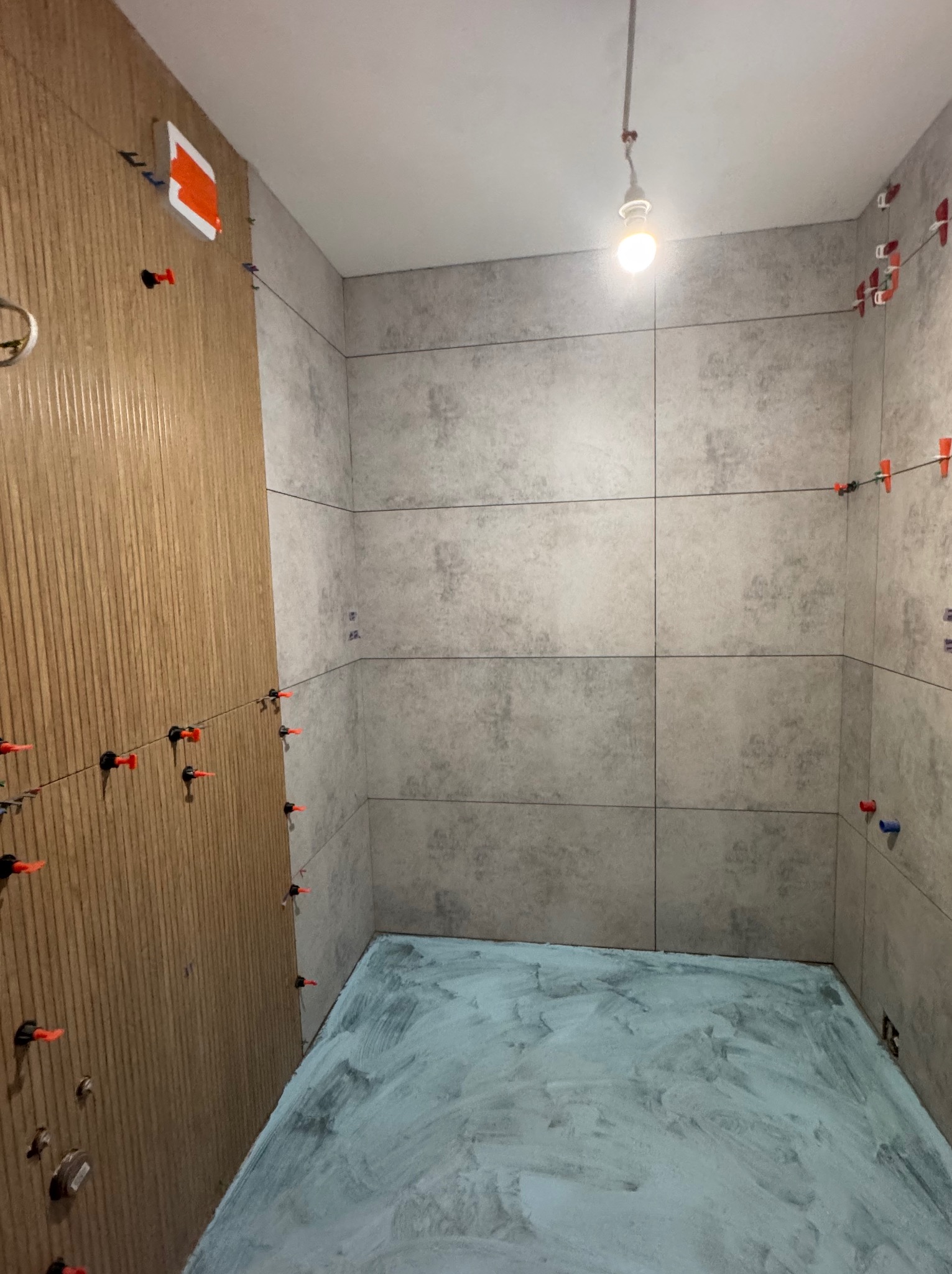 Proces układania płytek w łazience: szare płytki ścienne z systemem poziomowania, przygotowane podłoże podłogowe w trakcie prac glazurniczych, surowa żarówka.