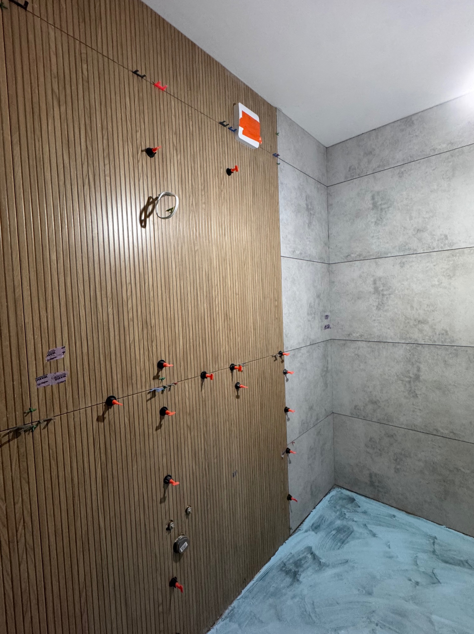 Montaż płytek w łazience: brązowe panele ścienne z klipsami poziomującymi, szare płytki na sąsiedniej ścianie, podłoga pokryta klejem.