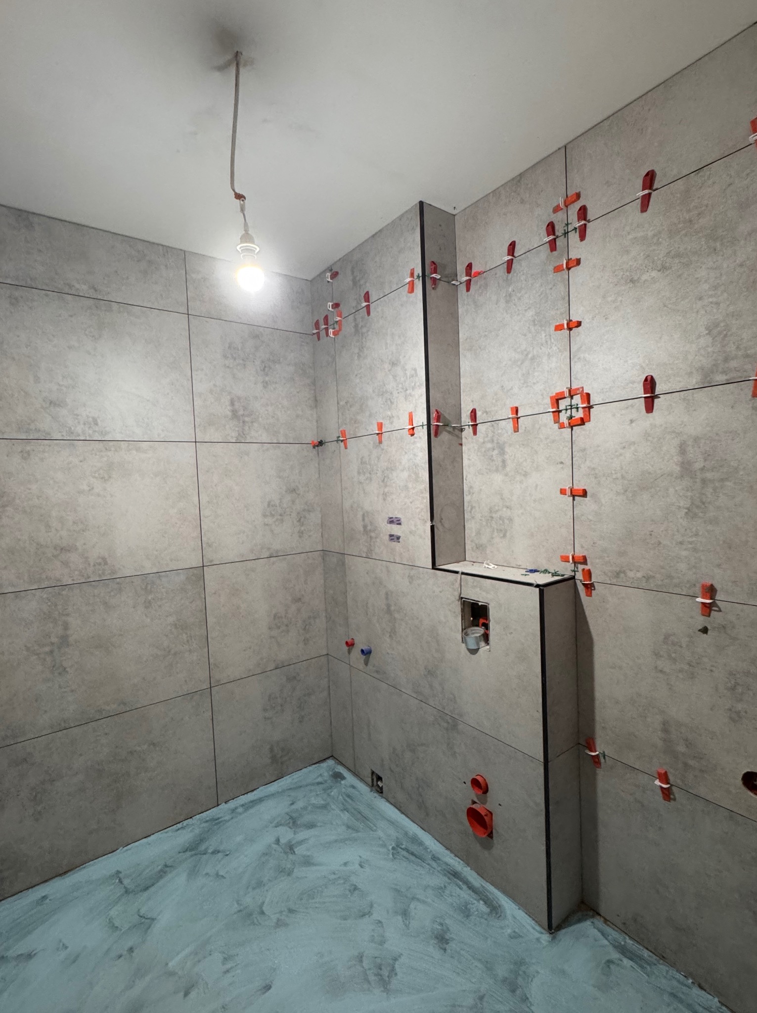 Szare płytki w trakcie układania na ścianach łazienki, z użyciem czerwonych krzyżyków dystansowych. Podłoga pokryta warstwą hydroizolacji, widoczne elementy instalacji.