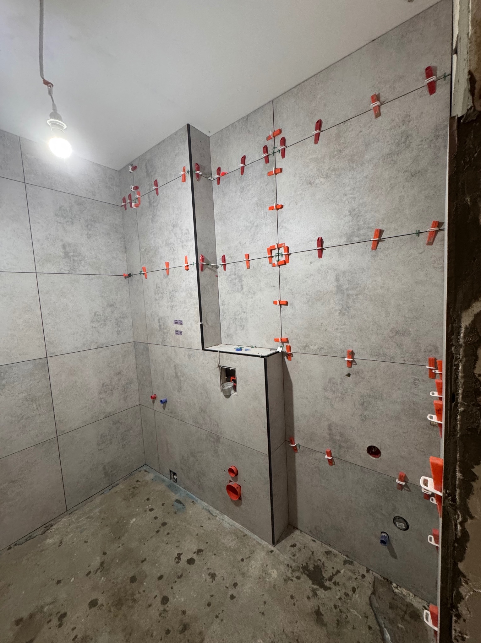 Szare płytki w łazience, w trakcie układania, z użyciem poziomicy i klipsów. Widoczne otwory pod instalacje, surowa podłoga i niedokończone wykończenie narożnika.