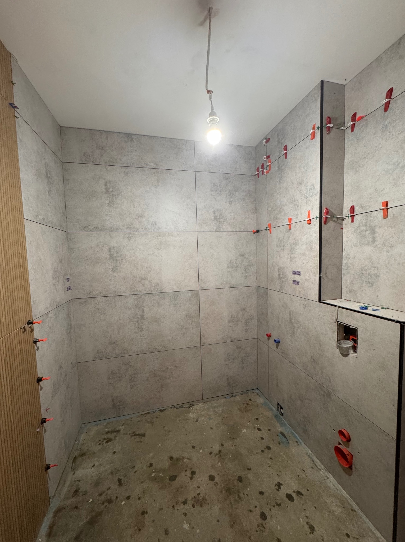 Wnętrze łazienki w trakcie prac glazurniczych. Szare płytki ułożone na ścianach z użyciem klipsów poziomujących. Częściowo wykończona ściana z drewnopodobnych paneli.