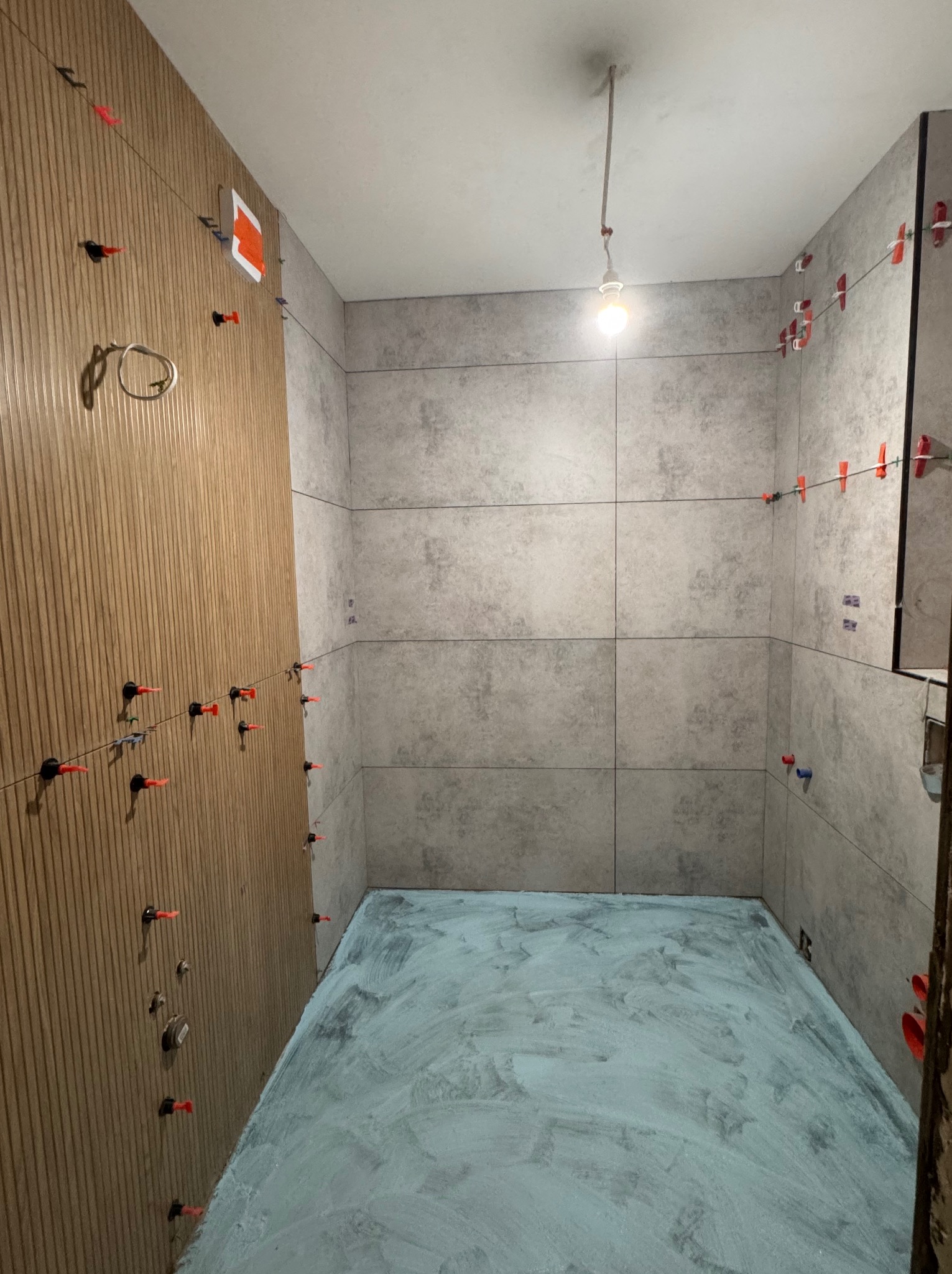 Łazienka w trakcie prac glazurniczych: szare płytki na ścianach z systemem poziomowania, przygotowane podłoże na podłodze, drewnopodobna ściana z instalacją.