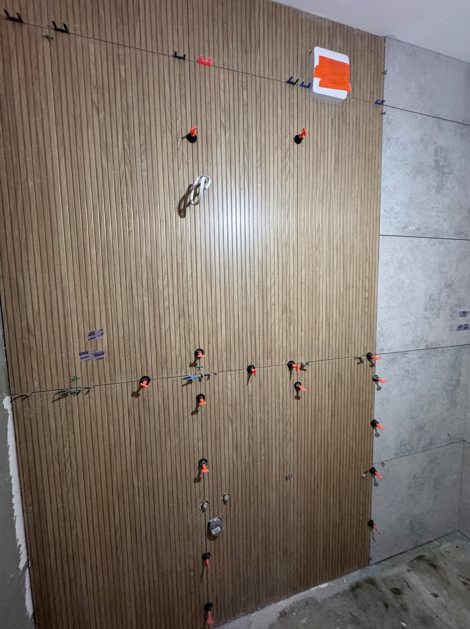 Montaż paneli imitujących drewno na ścianie, z użyciem poziomicy laserowej i klipsów dystansowych. Częściowo ułożone płytki w szarym odcieniu.