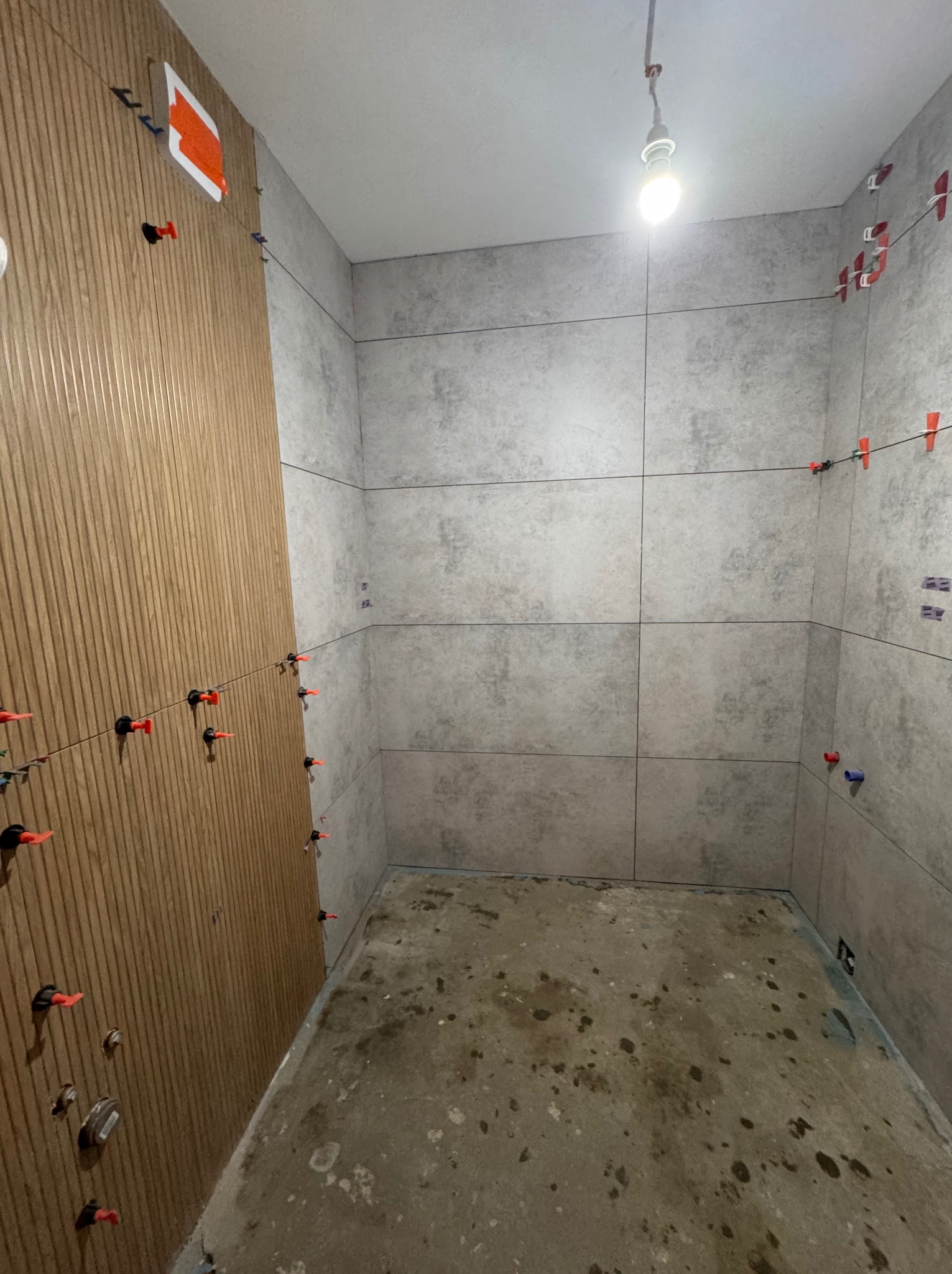 Wnętrze łazienki w trakcie prac glazurniczych: częściowo ułożone szare płytki na ścianach i dekoracyjna okładzina drewnopodobna. Widoczne systemy poziomowania płytek.