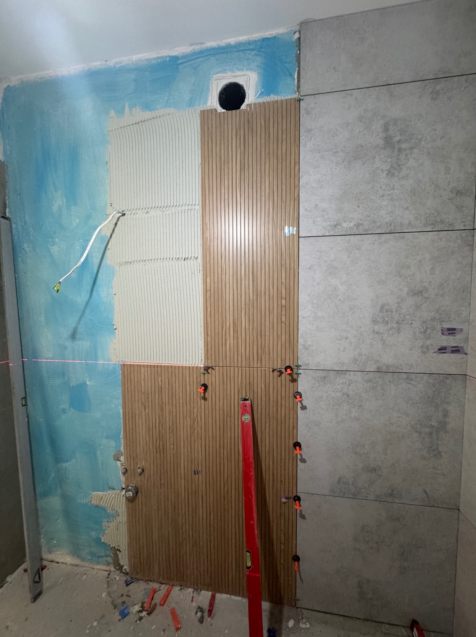 Montaż okładziny ściennej: część ściany pokryta klejem, fragment z panelami dekoracyjnymi oraz ułożone duże płytki w szarym odcieniu. Poziomica pionuje panele.