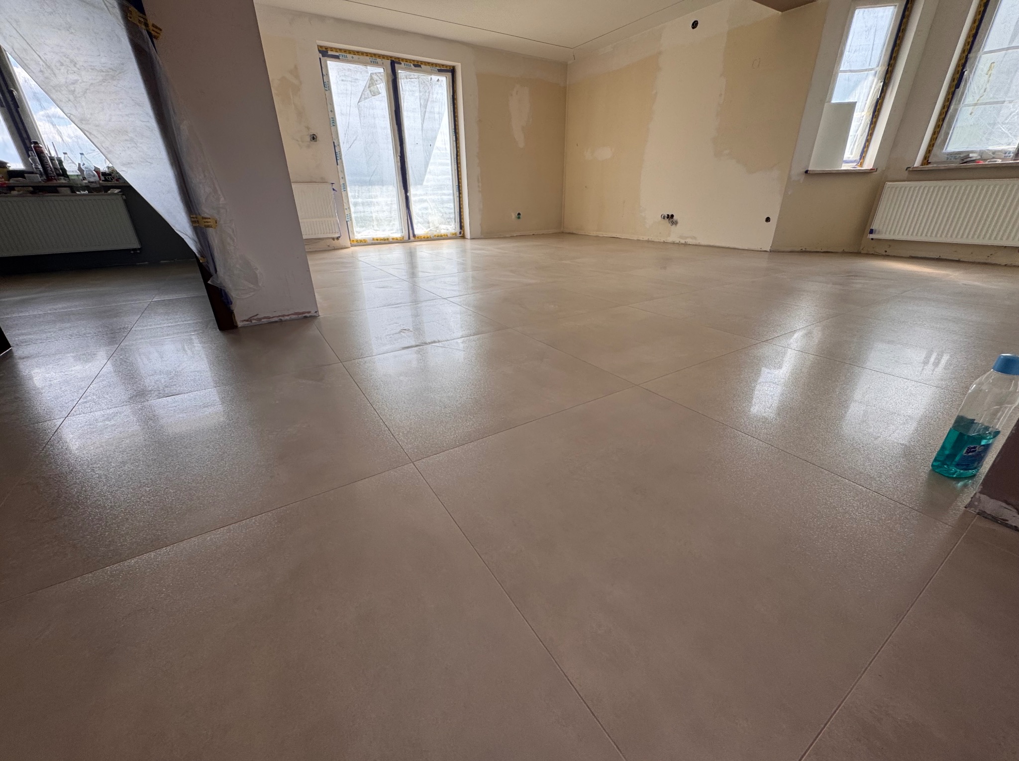 Wykończona podłoga z płytek w przestronnym, jasnym wnętrzu. Ściany przygotowane do malowania, okna zabezpieczone folią. Widoczna butelka płynu do czyszczenia.