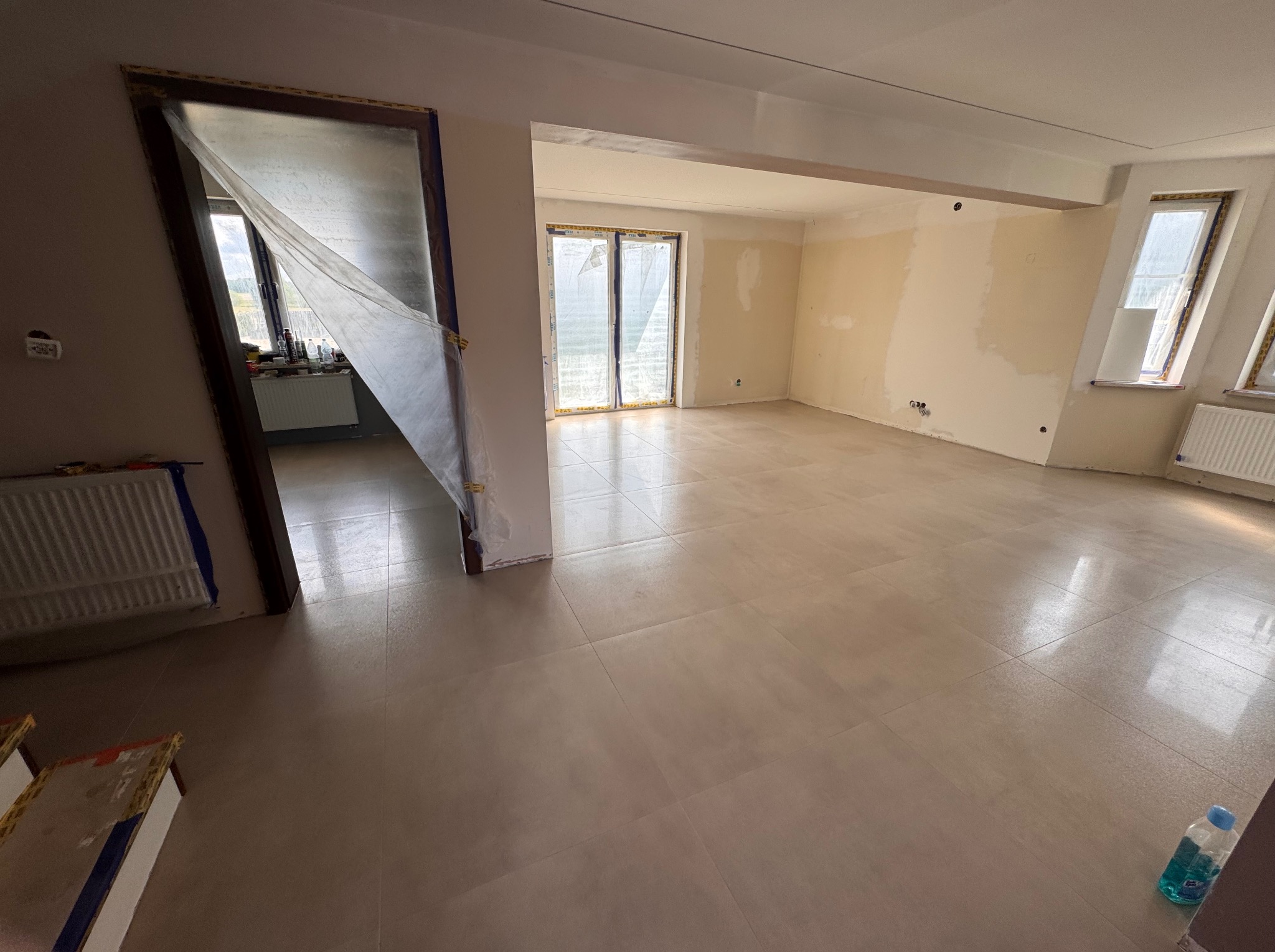 Wykończone płytki podłogowe w jasnym odcieniu beżu w przestronnym, częściowo odnowionym wnętrzu. Zabezpieczone drzwi i okna folią malarską.