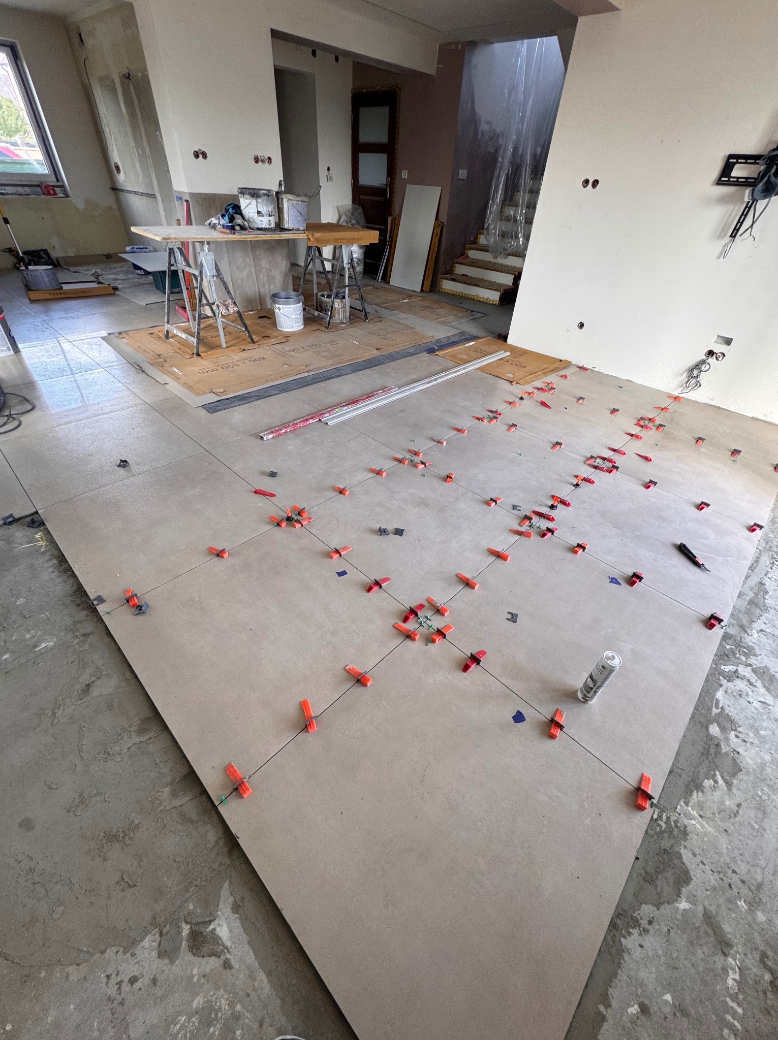 Układanie beżowych płytek podłogowych z użyciem klipsów poziomujących w przestronnym, jasnym wnętrzu. Widoczne narzędzia i przygotowane podłoże.