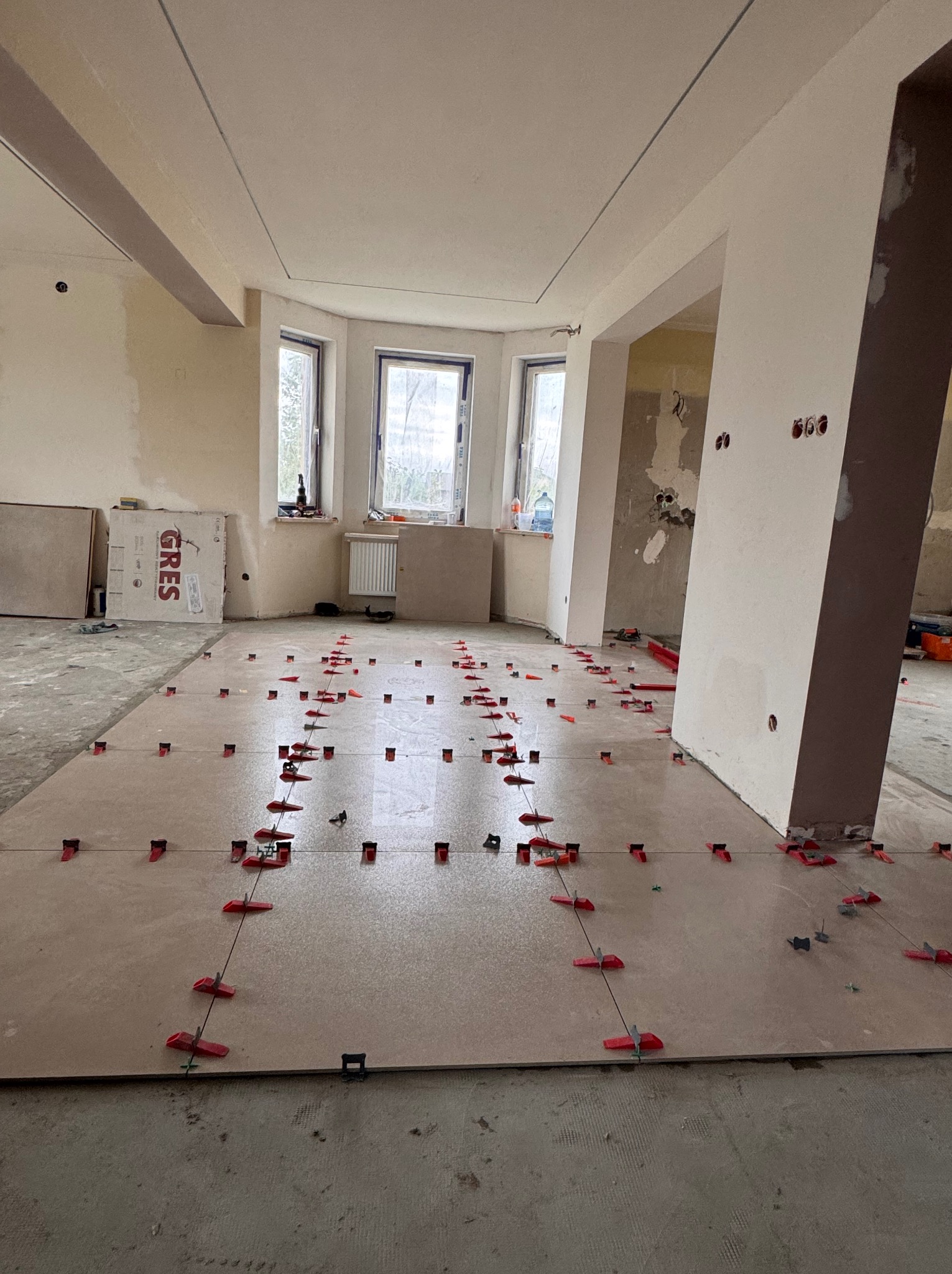 Układanie płytek podłogowych z użyciem klipsów poziomujących w przestronnym, jasnym wnętrzu. Gres na podłodze, przygotowany do fugowania.