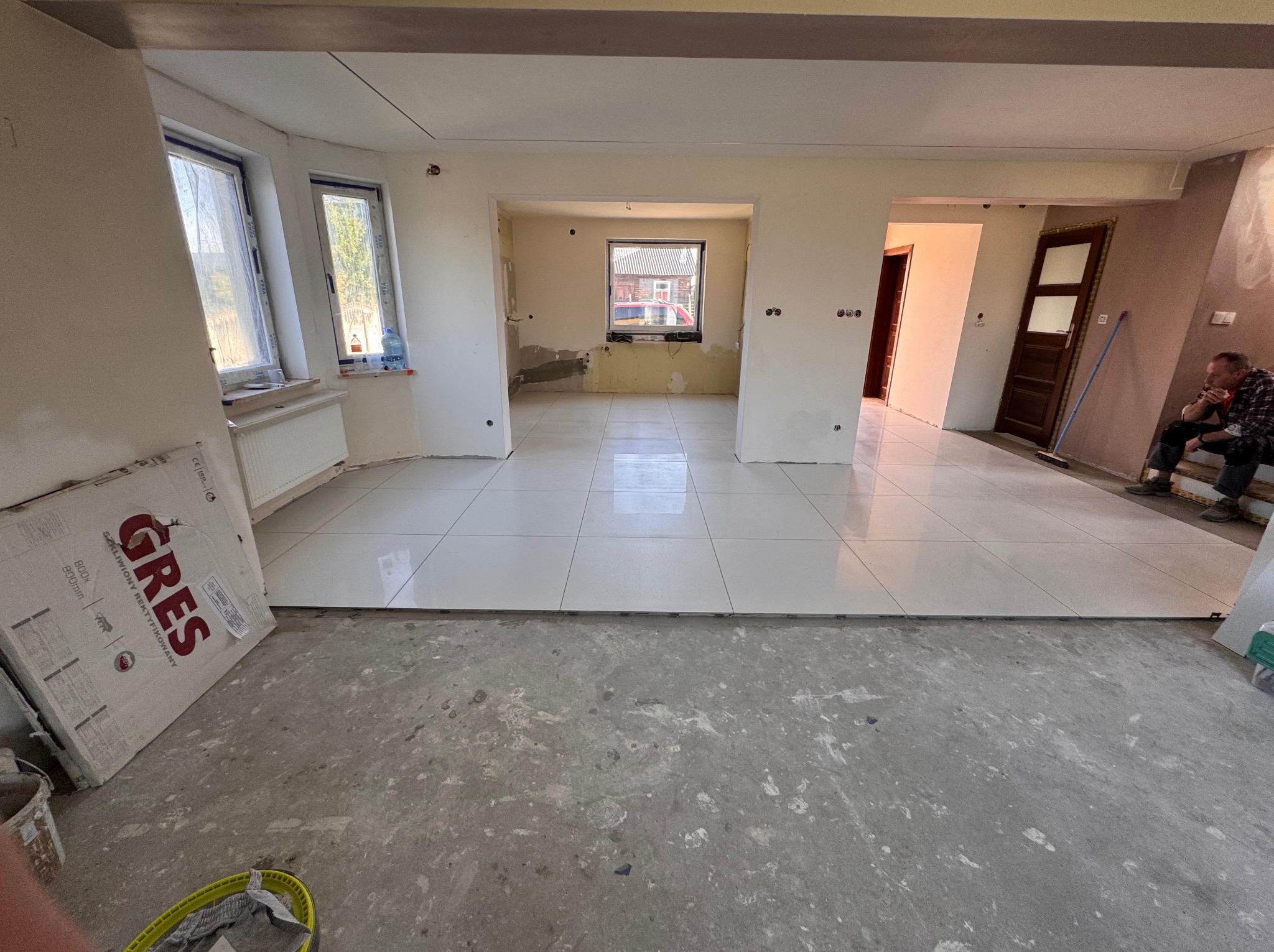 Wnętrze w trakcie układania białych płytek gresowych na podłodze. Widoczne okna, grzejnik, karton z napisem 'GRES' i siedząca osoba w oddali.