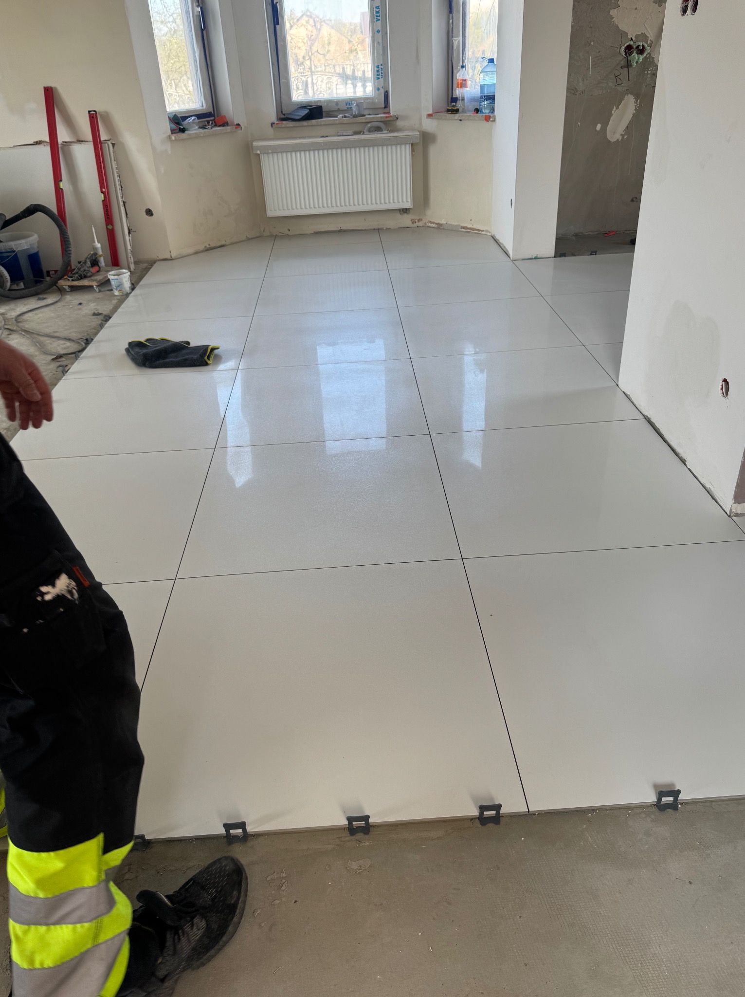 Błyszczące, białe płytki podłogowe w trakcie układania, z widocznymi elementami poziomującymi i fragmentem stroju roboczego glazurnika. Jasne wnętrze w stanie surowym.